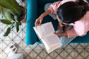A woman sitting in a cozy armchair reading a physical book, illustrating phone free activities at home. ©nenetus