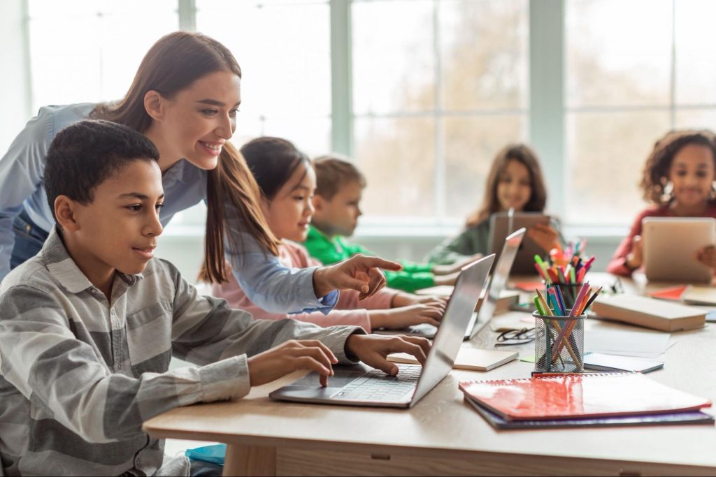 A teacher helps young elementary students use laptops in a bright classroom, representing top-rated Forsyth County Schools like Pooles Mill Elementary. ©Prostock-studio