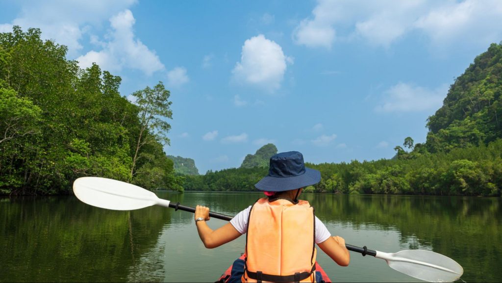 A person wearing a sun hat and life vest kayaks down a calm river surrounded by green trees, representing the Etowah River access at Etowah Landing. ©Porstocker